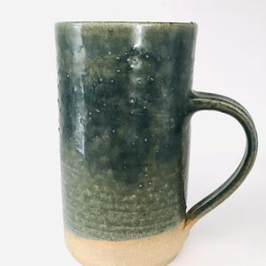 May include: A tall cylindrical ceramic mug featuring a dark forest green glaze with a mottled, textured finish on the upper portion and a smooth, unglazed beige clay base. The mug includes a curved handle attached to the side of the vessel.