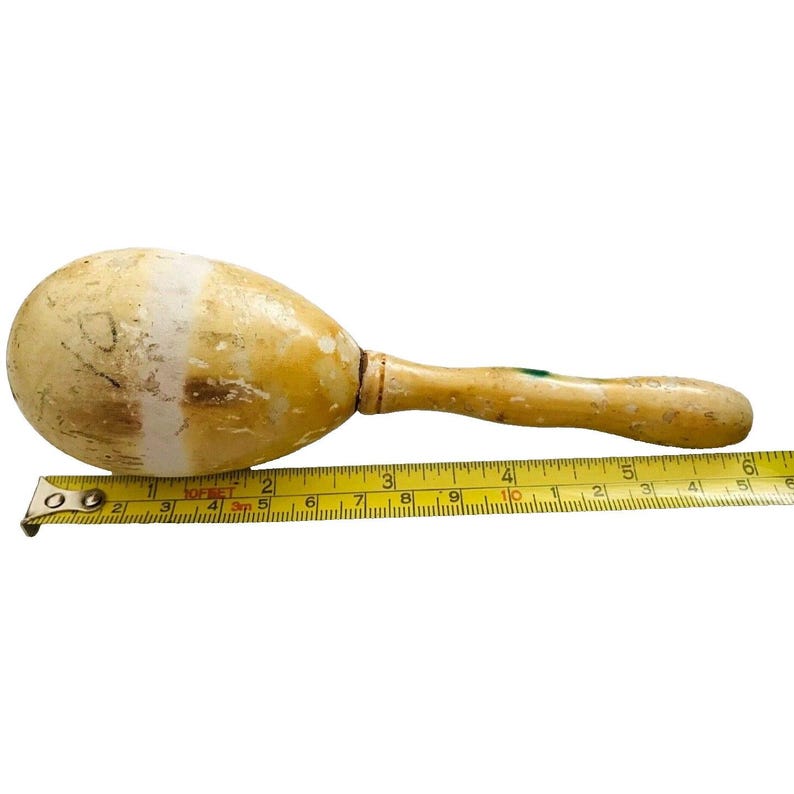 Puede incluir: Una maraca vintage con un mango marr&oacute;n claro y una cabeza en forma de huevo de color crema. La cabeza mide aproximadamente 10 cm de largo. Una cinta m&eacute;trica amarilla se coloca junto a la maraca.