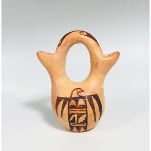Puede incluir: Un pequeño recipiente de cerámica de color beige claro con una forma distintiva, con una abertura central y dos picos laterales. El frente está decorado con un diseño de pájaro marrón oscuro, mostrando el arte nativo americano. El recipiente es probablemente hecho a mano.