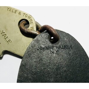May include: Close-up of two vintage metal keychains. One keychain is a light beige color with the text "YALE & TOWNE MFG" and "YALE". The other keychain is dark gray with the text "JOHNNIE WALKER N.Y."