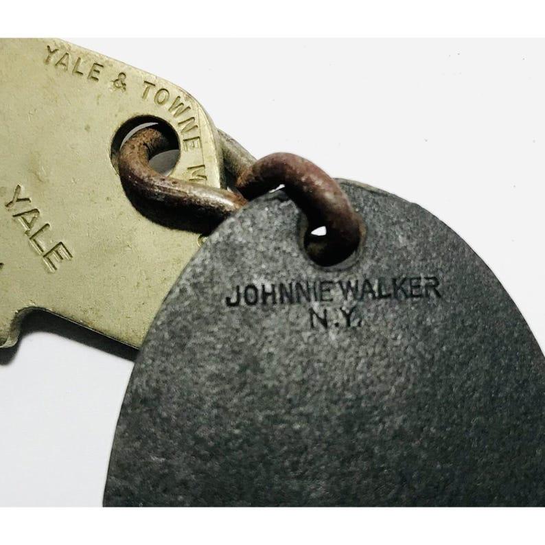 May include: Close-up of two vintage metal keychains. One keychain is a light beige color with the text "YALE & TOWNE MFG" and "YALE". The other keychain is dark gray with the text "JOHNNIE WALKER N.Y."