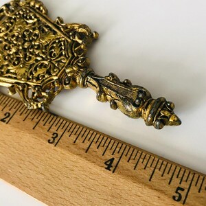 May include: A gold-coloured metal key with intricate details and a decorative handle. The key is approximately 10 cm long and is measured against a wooden ruler.