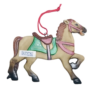 May include: A wooden carousel horse ornament with a green and pink saddle, red string hanger, and a "Schmid" label.