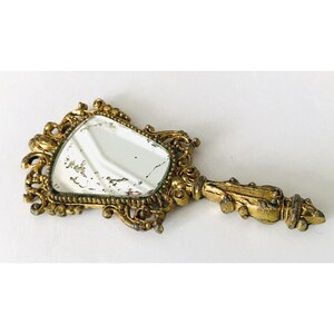 May include: A gold-toned hand mirror with an ornate, intricate design. The mirror has a slightly oval shape and is surrounded by a decorative frame.