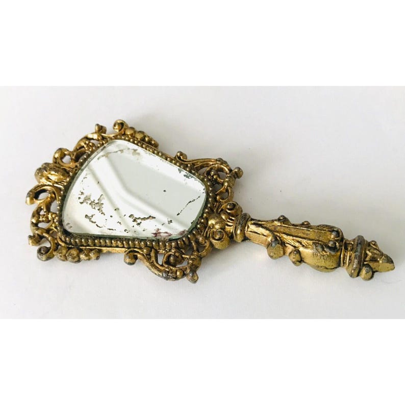 May include: A gold-toned hand mirror with an ornate, intricate design. The mirror has a slightly oval shape and is surrounded by a decorative frame.