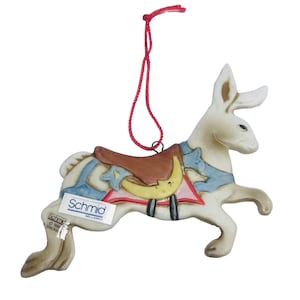 Könnte beinhalten: Weißer Porzellan-Hasen-Anhänger mit blauem und braunem Sattel, einer roten Schnur zum Aufhängen und dem Text "Schmid Sri Lanka" auf der Unterseite.