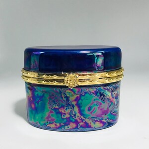 May include: A small, round, iridescent trinket box with a dark blue lid and a gold-colored band. The box has a swirling pattern of blues, greens, and purples. The band is decorated with a floral clasp.