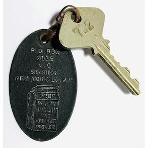 May include: Vintage key and key tag. The key is silver with the number 712. The tag is oval-shaped, dark gray, and has text that reads "P.O. BOX 1115, S.C. STATION, NEW YORK 17, NY" and "DROP IN ANY MAILBOX WE GUARANTEE POSTAGE."