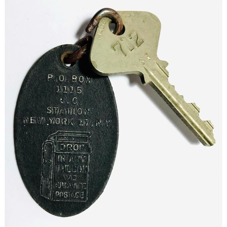 May include: Vintage key and key tag. The key is silver with the number 712. The tag is oval-shaped, dark gray, and has text that reads "P.O. BOX 1115, S.C. STATION, NEW YORK 17, NY" and "DROP IN ANY MAILBOX WE GUARANTEE POSTAGE."