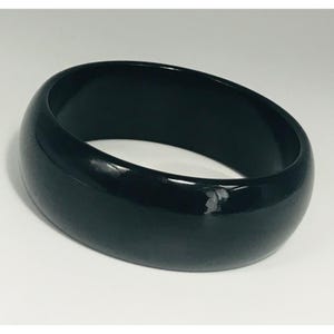May include: A glossy, black bangle bracelet. The wide, rounded band has a smooth surface and a slight sheen. The bracelet is a solid, dark color and appears to be made of a hard material.
