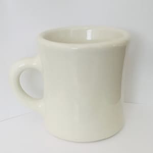 May include: A solid off-white ceramic mug with a thick, rounded handle and a smooth, glossy finish. The mug features a cylindrical body with a slightly flared rim and a sturdy base, presenting a simple, functional design for holding hot beverages.