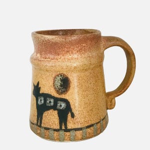 May include: A ceramic mug with a speckled tan and brown glaze. The mug features a dark silhouette of a four-legged animal with three small square patterns on its body, a dark circular shape above it, and a striped pattern along the base.