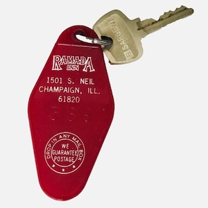 May include: A vintage red Ramada Inn key tag with a metal key attached. The tag has the Ramada Inn logo and address details: 1501 S. Neil, Champaign, IL, 61820. The key is silver and has the word "SARGENT" printed on it.