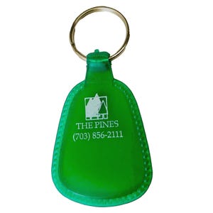 May include: A green plastic keyring with a gold-coloured ring. The keyring has a rounded shape with a beaded edge and features the text "THE PINES" and a phone number.