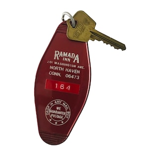 May include: A vintage maroon Ramada Inn key tag with a gold-coloured key attached. The tag has the Ramada Inn logo and address details, including "201 Washington Ave. North Haven, CONN. 06473." The key reads "Taylor 64."