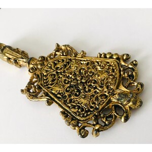 May include: A gold-toned metal key with intricate floral designs. The key has a decorative handle and a pointed end.