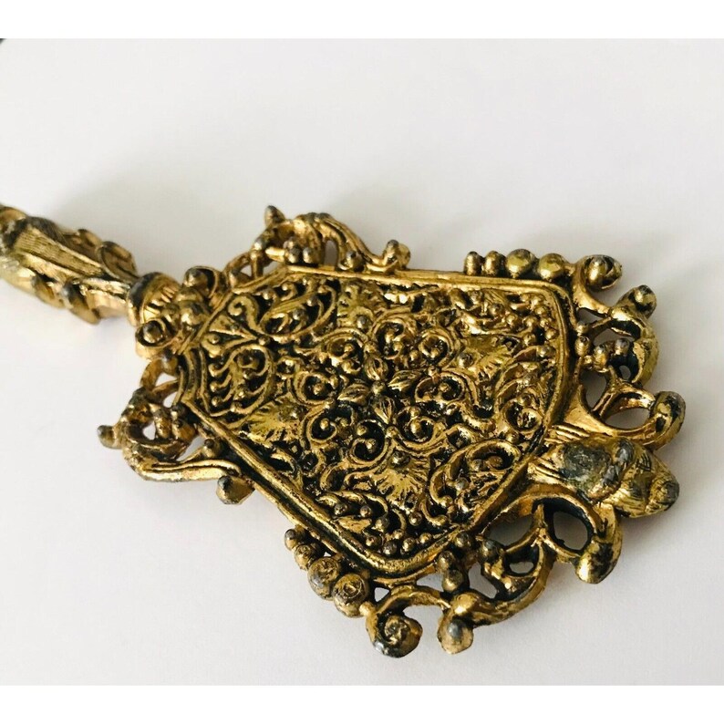 May include: A gold-toned metal key with intricate floral designs. The key has a decorative handle and a pointed end.