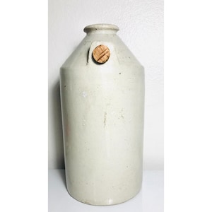 Może przedstawiać: Biała ceramiczna butelka z drewnianym korkiem. Butelka ma zaokrąglone ciało i wąską szyjkę. Korek jest prostym okrągłym dyskiem z wzorem słojów drewna.