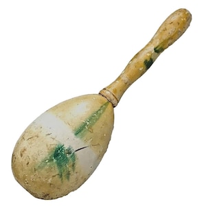 Puede incluir: Una maraca de madera vintage con un cuerpo y mango de color beige claro. El cuerpo tiene detalles en blanco y verde. El mango est&aacute; en &aacute;ngulo y tiene un aspecto desgastado. La maraca es un instrumento musical.