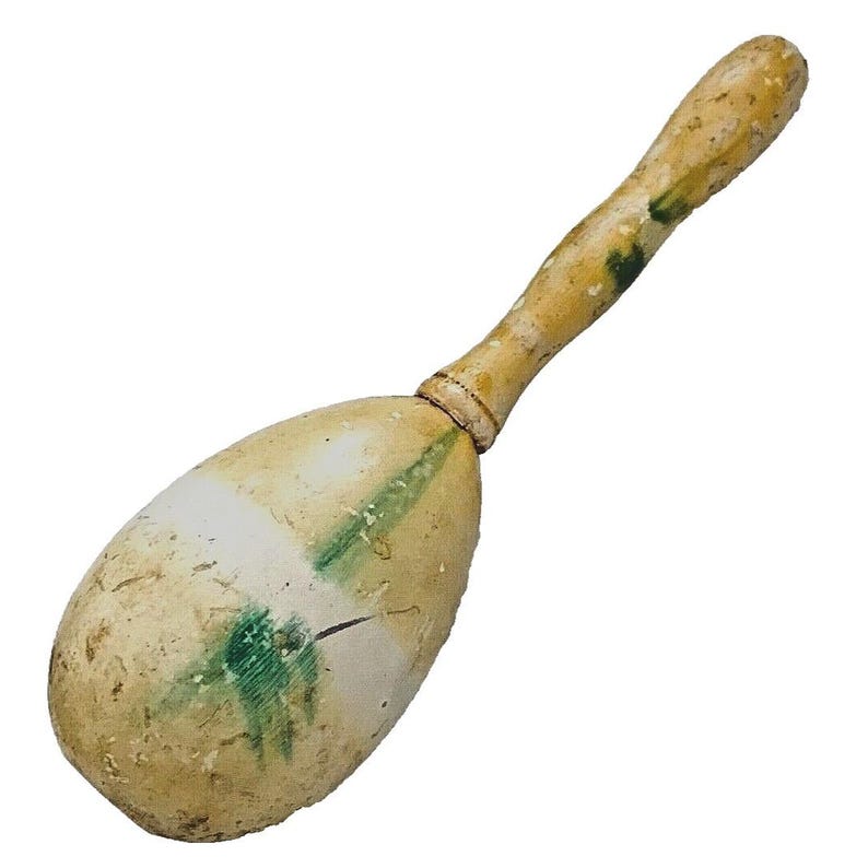 Puede incluir: Una maraca de madera vintage con un cuerpo y mango de color beige claro. El cuerpo tiene detalles en blanco y verde. El mango est&aacute; en &aacute;ngulo y tiene un aspecto desgastado. La maraca es un instrumento musical.