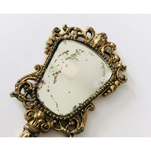 May include: A gold-toned hand mirror with an ornate, decorative frame. The mirror has a slightly tarnished, aged look.