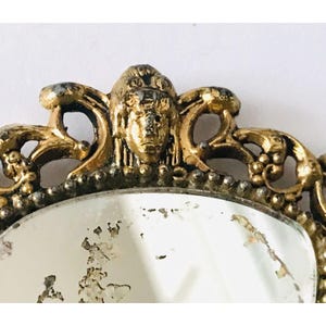May include: An ornate gold-toned frame with a round mirror. The frame features a decorative design with a central face and intricate scrollwork.