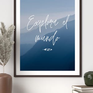 May include: Framed art print featuring a mountain landscape in shades of blue. The white cursive text reads "Explora el mundo" with a small leaf illustration. The artwork is displayed on a wooden shelf.