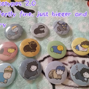 May include: A collection of eleven round buttons with cartoon character designs. The buttons are in various colors, including gray, pink, yellow, and blue. Text at the top reads "Prototypes 2.0 Perfectly fine, just bigger and blurry."