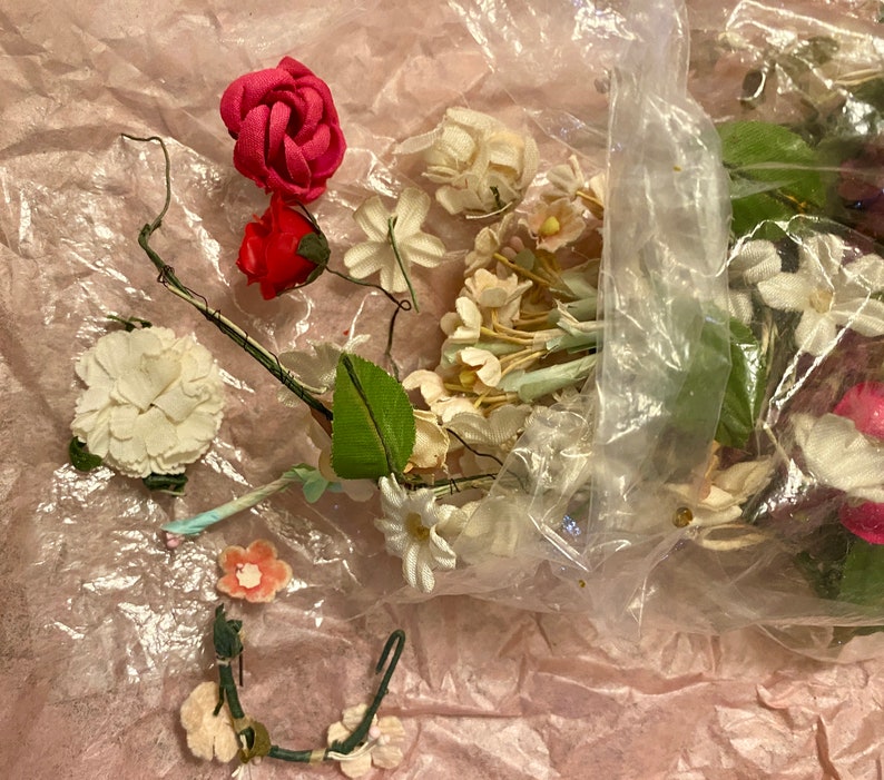 Op de afbeelding: Een plastic zak gevuld met verschillende kunstbloemen, waaronder roze rozen, witte madeliefjes en groene bladeren. De bloemen zijn losjes en natuurlijk gerangschikt, en de zak is open aan de bovenkant, waardoor de inhoud zichtbaar is.
