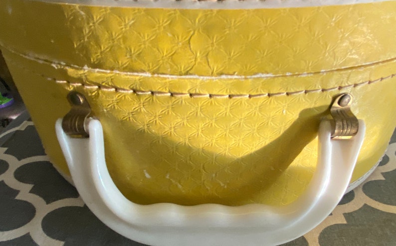 Op de afbeelding: Een gele vintage hoedenbox met een witte kunststof handgreep en gouden metalen sluitingen. De doos heeft een getextureerd oppervlak met een herhalend patroon.
