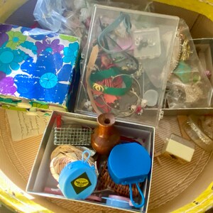 Op de afbeelding: Een gele rieten mand gevuld met verschillende kleine dozen en containers. De dozen bevatten een verscheidenheid aan items, waaronder sieraden, een houten flessenstop en een blauwe plastic container. De mand is gevuld met verschillende items, waaronder sieraden, een houten flessenstop en een blauwe plastic container.