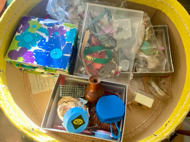 Op de afbeelding: Een gele rieten mand gevuld met verschillende kleine dozen en containers. De dozen bevatten een verscheidenheid aan items, waaronder sieraden, een houten flessenstop en een blauwe plastic container. De mand is gevuld met verschillende items, waaronder sieraden, een houten flessenstop en een blauwe plastic container.