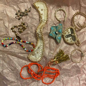 Op de afbeelding: Een verzameling sieraden en accessoires met verschillende stijlen en kleuren. De items omvatten een kralenketting, een parelketting, een kralenarmband, een paar oorbellen, een klein handtasje en een decoratief stuk met parels en pailletten.