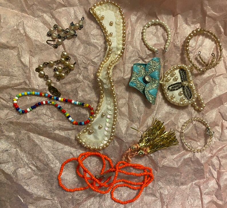 Op de afbeelding: Een verzameling sieraden en accessoires met verschillende stijlen en kleuren. De items omvatten een kralenketting, een parelketting, een kralenarmband, een paar oorbellen, een klein handtasje en een decoratief stuk met parels en pailletten.
