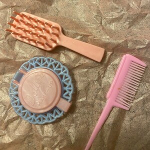 Op de afbeelding: Een set van drie roze plastic poppenaccessoires: een haarborstel, een kam en een compact met een blauw en wit kantmotief. De compact heeft een roze deksel met de tekst "Doll Make-Up" erop gedrukt.