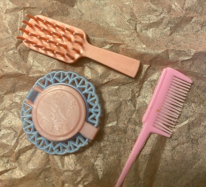 Op de afbeelding: Een set van drie roze plastic poppenaccessoires: een haarborstel, een kam en een compact met een blauw en wit kantmotief. De compact heeft een roze deksel met de tekst "Doll Make-Up" erop gedrukt.