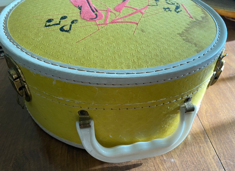 Op de afbeelding: Een ronde hoedendoos in geel met een witte handgreep en een witte rand rond de bovenkant. Het deksel heeft een roze en rood ontwerp met muzieknoten en de woorden "Let's Have Fun".
