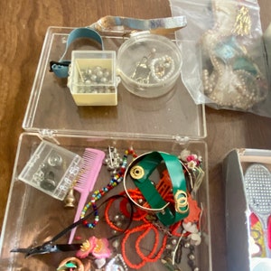 Op de afbeelding: Een doorzichtige plastic container gevuld met diverse sieraden en snuisterijen, waaronder kralen, een kam, een klein doosje parels, een groene armband en een cassettebandje.