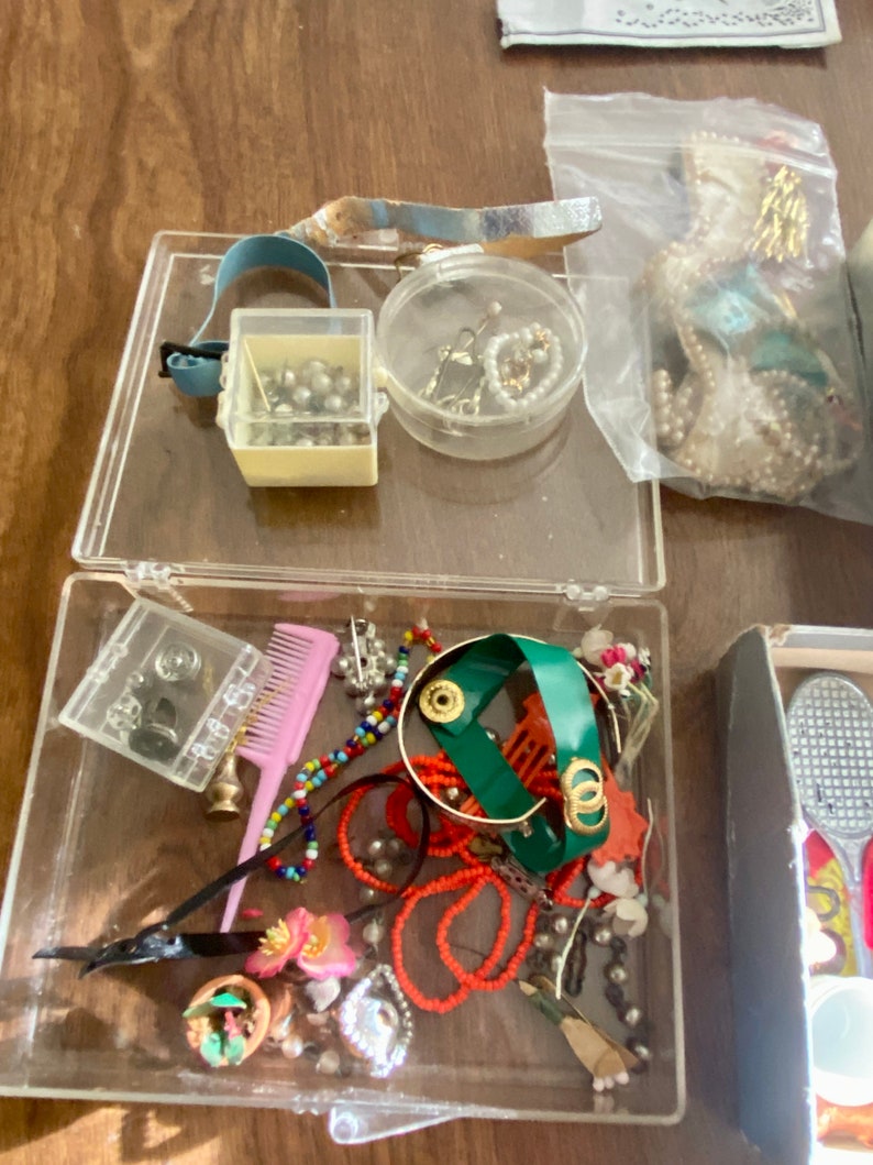 Op de afbeelding: Een doorzichtige plastic container gevuld met diverse sieraden en snuisterijen, waaronder kralen, een kam, een klein doosje parels, een groene armband en een cassettebandje.