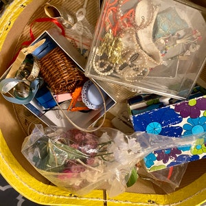 Op de afbeelding: Een gele vintage koffer gevuld met verschillende items, waaronder een rieten mand, een doorzichtige plastic doos met sieraden, een plastic zak met gedroogde bloemen en een blauw en groen boek met een patroon.