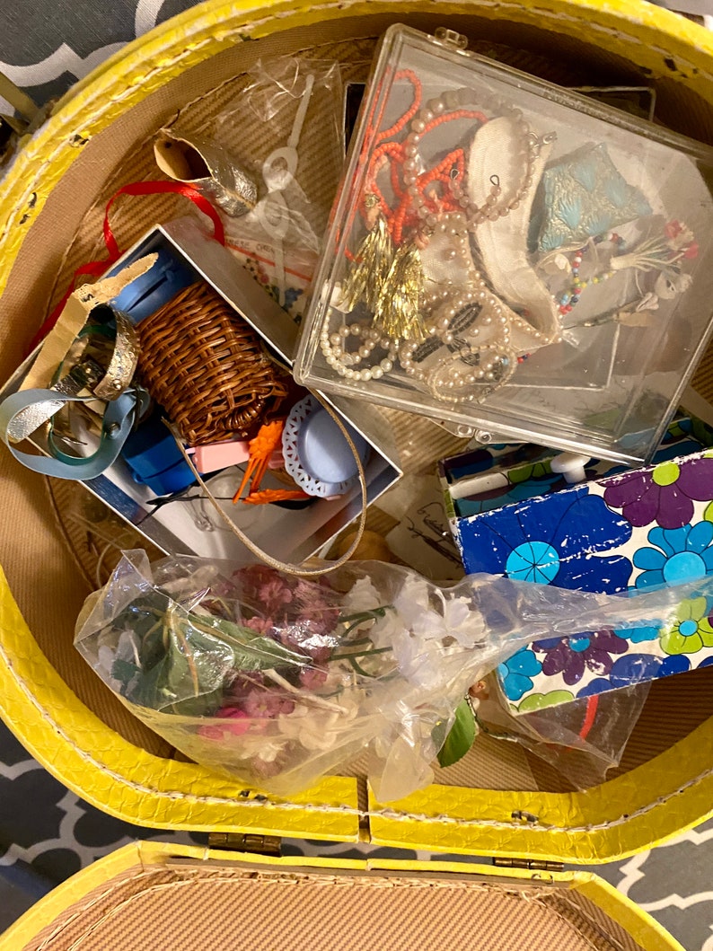 Op de afbeelding: Een gele vintage koffer gevuld met verschillende items, waaronder een rieten mand, een doorzichtige plastic doos met sieraden, een plastic zak met gedroogde bloemen en een blauw en groen boek met een patroon.