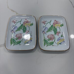 May include: Two square, white ceramic dishes with rounded corners. Each dish features a floral design in pink, blue, yellow, and green, with gold trim. The floral pattern includes leaves and vines. The dishes are displayed on a white surface.