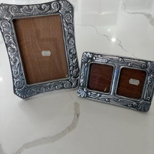 May include: Two ornate silver-toned picture frames. One is rectangular, the other has two square openings. Both frames have detailed floral and shell designs around the edges and wooden backing.