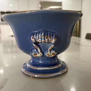 May include: A blue ceramic bowl with a flared rim and a pedestal base. The bowl features decorative raised accents in a lighter color with brown trim. The bowl is likely used for decorative purposes.