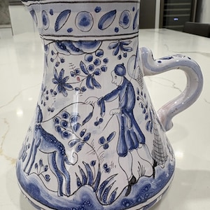 May include: A large, hand-painted ceramic pitcher with a blue and white design. The pitcher features a detailed illustration of figures and foliage. The handle is curved and painted with blue and pink accents. The base and rim have a blue border.