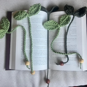 May include: Two crochet leaf bookmarks in a book. The leaves are green, with a braided green and brown cord. Wooden beads are attached to the ends of the cords. The book is open, with text visible on the pages.