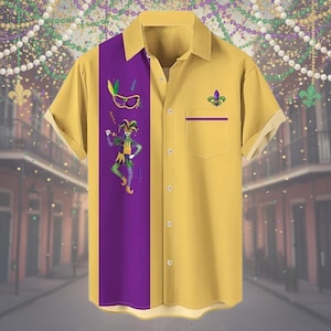 Könnte beinhalten: Gelbes Button-Down-Hemd mit einem vertikalen lila Panel mit einer Narren-Illustration und einer Mardi-Gras-Maske. Das Hemd hat eine Tasche mit lila Besatz und einem Fleur-de-Lis-Emblem. Das kurzärmelige Hemd ist ein festliches Design.