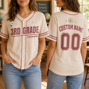 Könnte beinhalten: Ein cremefarbenes Baseballtrikot mit kastanienbraunen Streifen und Besatz. Die Vorderseite zeigt den Text 3RD GRADE und CUSTOM NAME mit einer Gänseblümchengrafik. Die Rückseite zeigt CUSTOM NAME und die Nummer 00 in Kastanienbraun mit einer Blume.