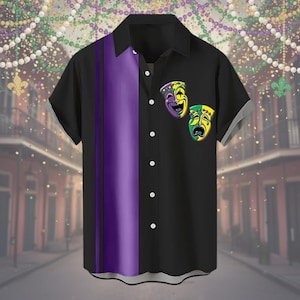 Può includere: Camicia nera a maniche corte con una striscia verticale viola e due grafiche di maschere teatrali. La camicia ha un colletto classico e bottoni bianchi. Le maschere sono gialle, verdi e viola.