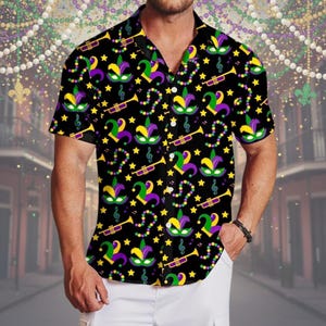 Könnte beinhalten: Schwarzes kurzärmeliges Hemd mit einem lebendigen Mardi-Gras-Muster. Das Design zeigt Narrenmasken in Lila, Grün und Gelb, Noten, Trompeten und Sterne. Das Hemd wird mit einer weißen Hose getragen.